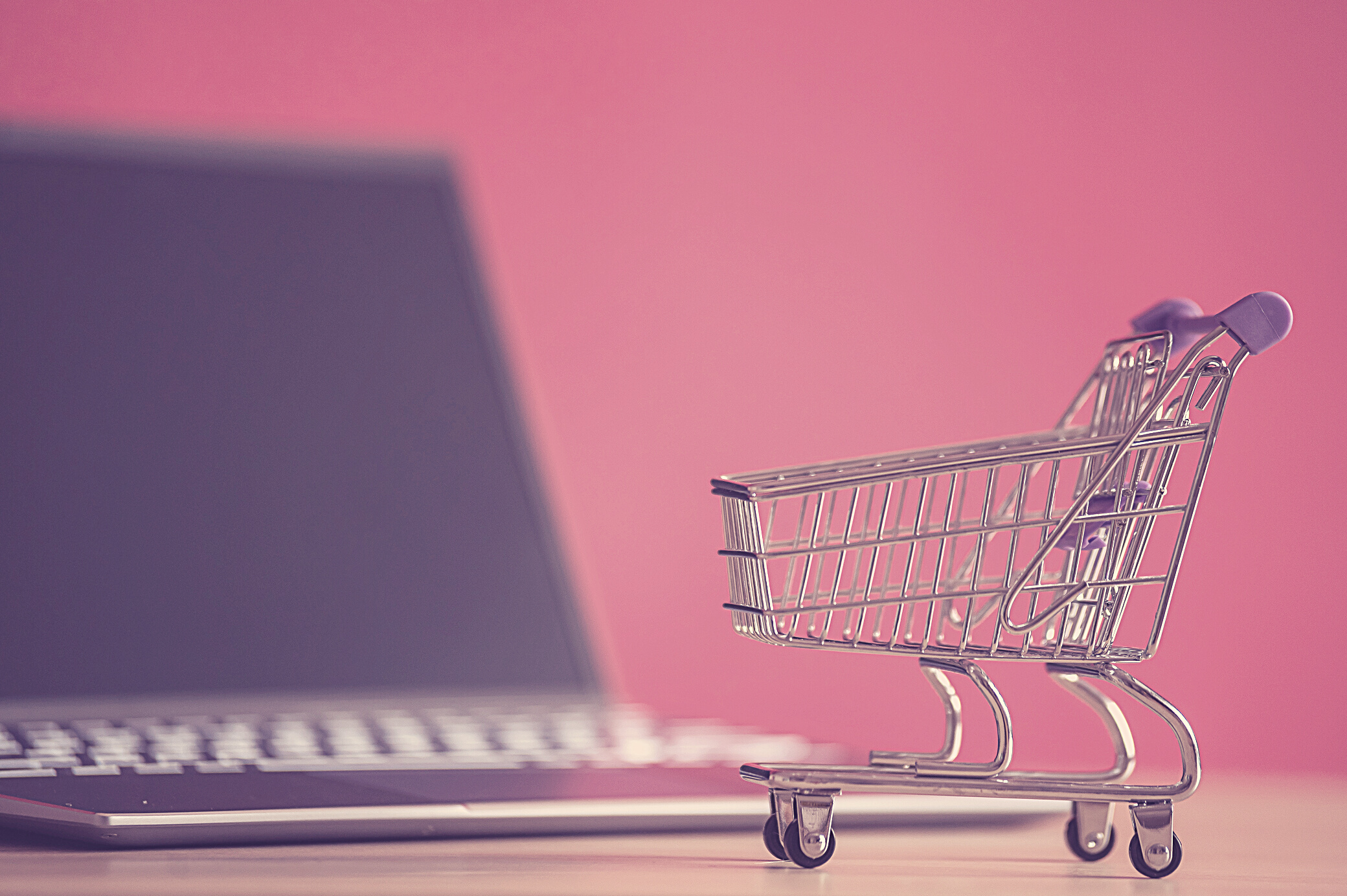 Online shopping concept. Mini grocery cart and laptop on a pink background. Small cart and personal computer